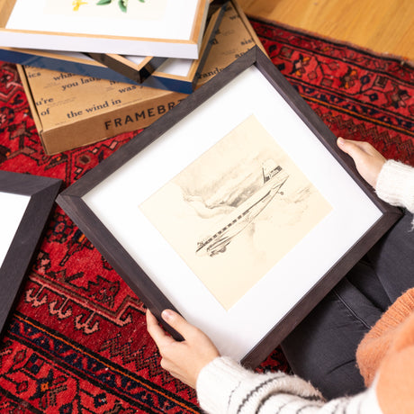 Woman holding framed drawing of airplane surrounded by framed art pieces on red rug on floor.