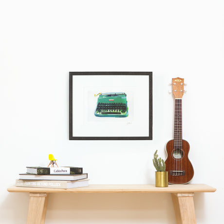 Green typewriter art in riveted black frame above table with ukulele cactus and books